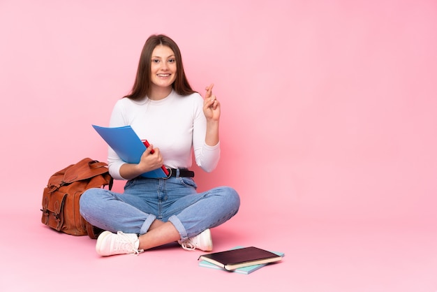 Adolescent caucasien étudiant fille assise sur le sol isolé sur rose avec les doigts traversant et souhaitant le meilleur