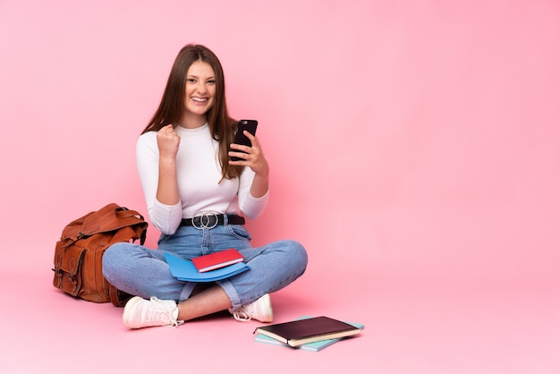 Adolescent caucasien étudiant fille assise sur le sol isolé sur rose avec téléphone en position de victoire