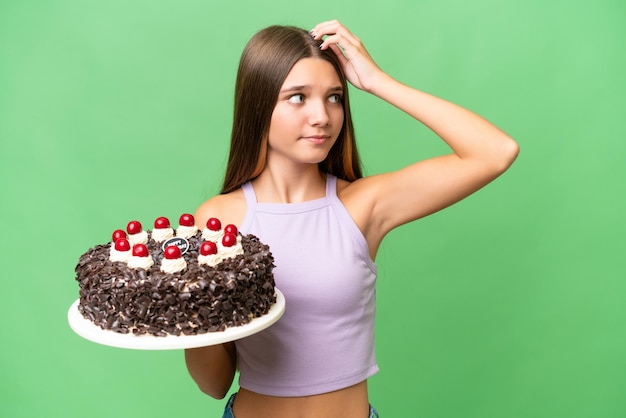 Adolescent caucasien fille tenant un gâteau d'anniversaire sur fond isolé ayant des doutes et avec une expression de visage confuse