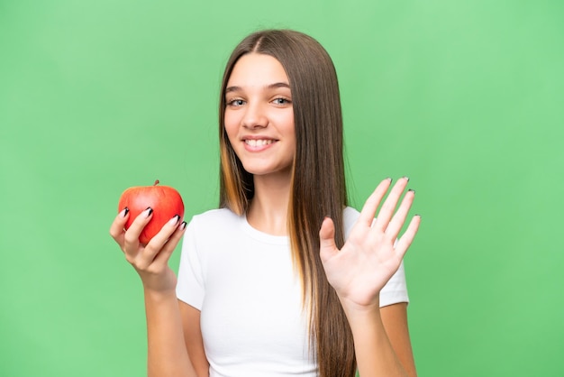 Adolescent caucasien fille tenant une pomme sur fond isolé saluant avec la main avec une expression heureuse