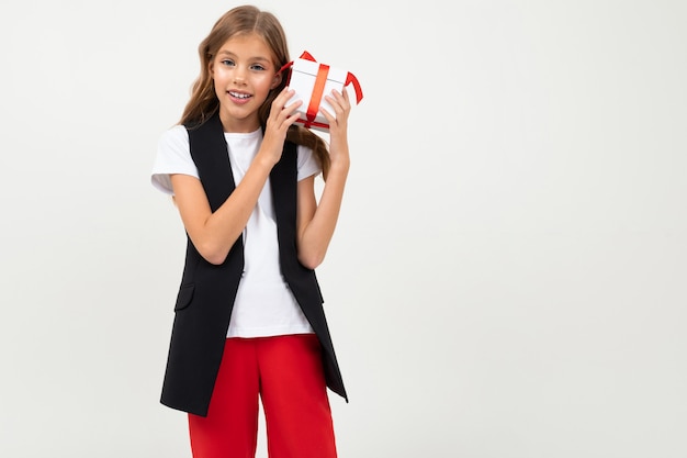 Adolescent caucasien fille en veste noire, chemise blanche et pantalon rouge est titulaire d'une boîte blanche avec cadeau et sourires isolé sur blanc