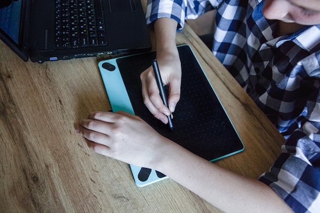 Un adolescent en chemise à carreaux dans le processus de formation à domicile dessine à l'aide d'une tablette graphique posée sur une table en bois.