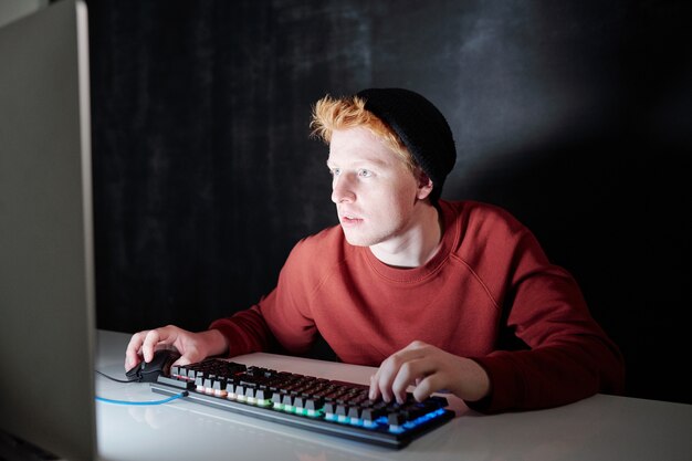 Adolescent contemporain en sweat-shirt et bonnet noir en appuyant sur les touches du clavier et en cliquant sur la souris alors qu'il était assis en face de l'écran de l'ordinateur