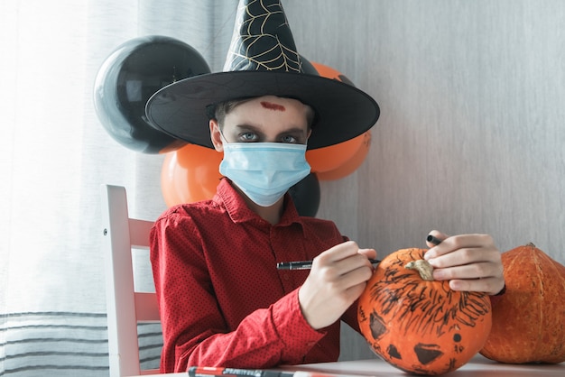 Adolescent en costume et masques pour se protéger du COVID-19 dessinant une citrouille pour la célébration d'Halloween