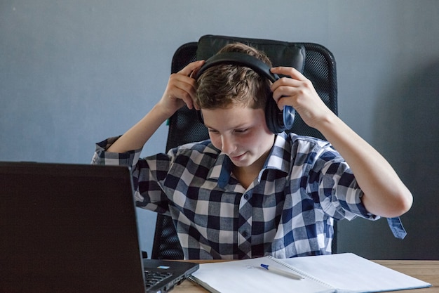 Un adolescent dans une chemise à carreaux étudie à la maison sur un ordinateur portable. Il est assis à la table avec un cahier ouvert dessus. Les écouteurs sont accrochés à l'ordinateur portable. Thème de l'enseignement à distance