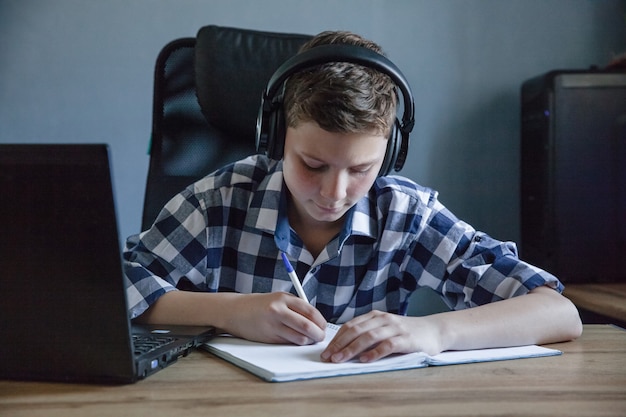 Un adolescent dans une chemise à carreaux étudie à la maison sur un ordinateur portable. Il est assis à la table avec un cahier ouvert dessus. Les écouteurs sont accrochés à l'ordinateur portable. Thème de l'enseignement à distance