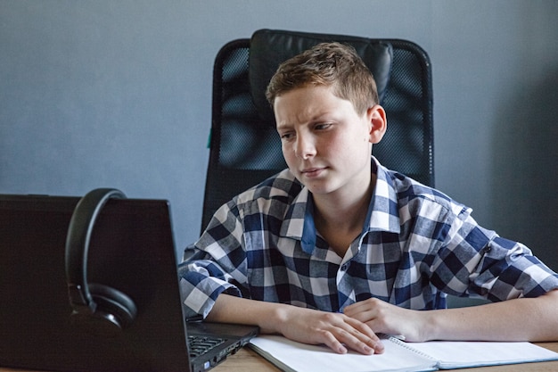 Un adolescent dans une chemise à carreaux étudie à la maison sur un ordinateur portable. Il est assis à la table avec un cahier ouvert dessus.