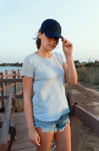 Photo adolescent debout au bord de la mer au coucher du soleil. teen girl wearing t-shirt et casquette de baseball bleu foncé et toucher la visière. visage à l'ombre. maquette de casquette et t-shirt