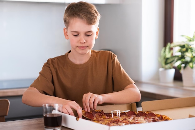 Un adolescent déchire un morceau de pizza dans la cuisine à la maison Livraison de nourriture