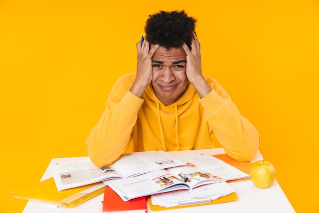 Adolescent désespéré étudiant assis au bureau avec des manuels isolés sur un mur jaune