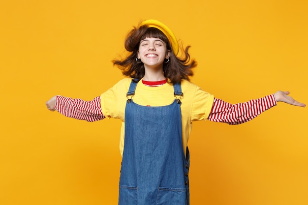 Adolescent drôle de fille en béret français, robe d'été en denim avec les yeux fermés, écartant les mains avec des cheveux flottants isolés sur fond jaune. Émotions sincères des gens, concept de style de vie. Maquette de l'espace de copie.