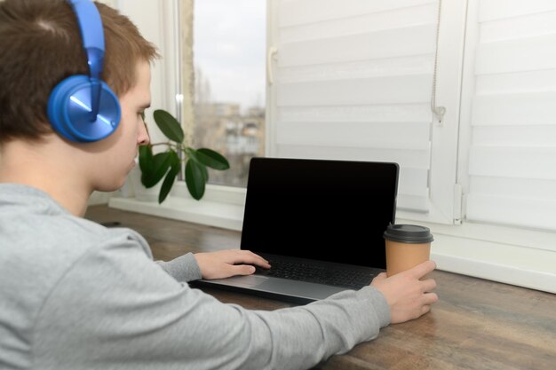 Un adolescent avec des écouteurs bleus est assis à l'ordinateur à la maison une tasse de café dans les mains