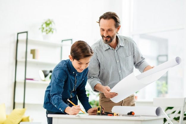 Adolescent enthousiaste émotionnel se penchant à la table tout en marquant la longueur de celui-ci à l'aide d'un ruban à mesurer et son père curieux debout avec un dessin près de