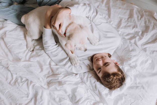 Un adolescent est allongé dans son lit sur une literie blanche avec un chien vue de dessus l'animal réveille le propriétaire le matin