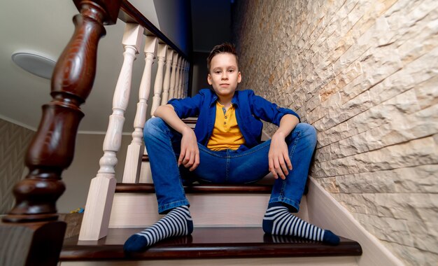 L'adolescent est assis détendu dans les escaliers. Escaliers en bois à la maison. Garçon regarde la caméra.