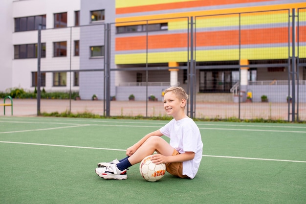 Un adolescent est assis sur un terrain vert dans la cour de l'école avec un ballon de football