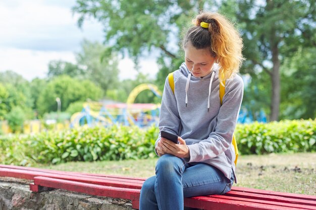 Adolescent étudiant fille avec sweat-shirt gris assis sur un banc dans le parc avec sac à dos et lecture de smartphone, espace copie