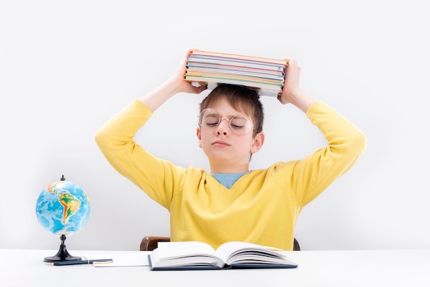 Adolescent fatigué assis au bureau et tenant une pile de cahiers au-dessus de sa tête Écolier difficile fait ses devoirs