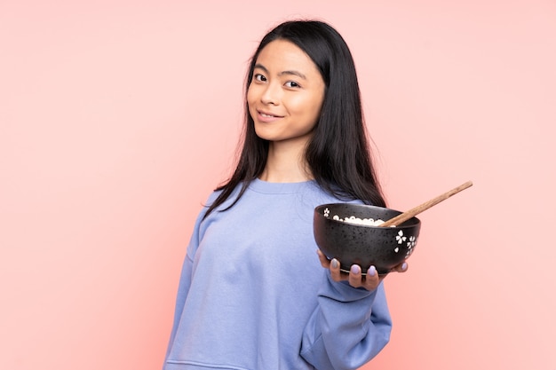 Adolescent femme asiatique sur mur beige avec une expression heureuse tout en tenant un bol de nouilles avec des baguettes
