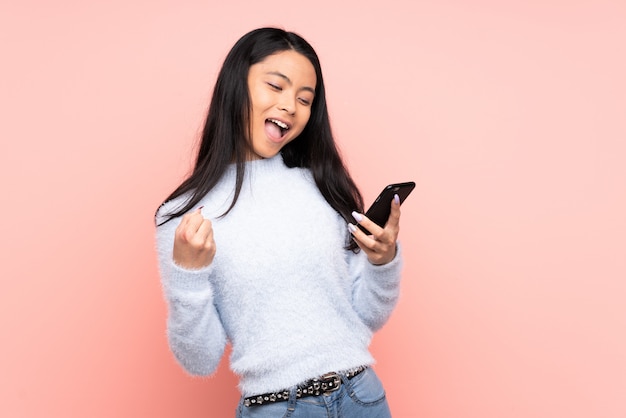 Adolescent femme chinoise isolée sur fond rose avec téléphone en position de victoire