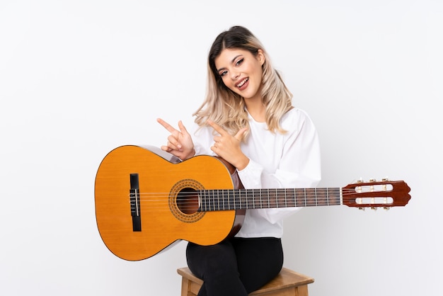 Adolescent femme avec guitare sur blanc isolé surpris et pointant le côté