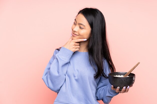 Adolescent femme isolé sur beige en pensant à une idée et à côté tout en tenant un bol de nouilles avec des baguettes