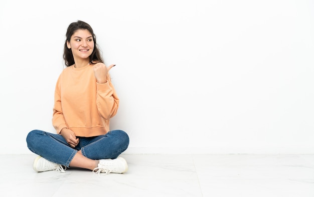 Adolescent femme russe assise sur le sol pointant vers le côté pour présenter un produit