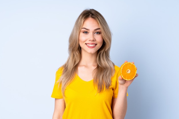 Adolescent femme tenant une orange isolée sur mur bleu souriant beaucoup