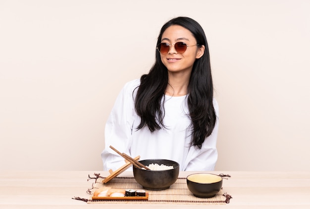 Adolescent fille asiatique manger de la nourriture asiatique isolé sur un mur beige avec des lunettes et heureux