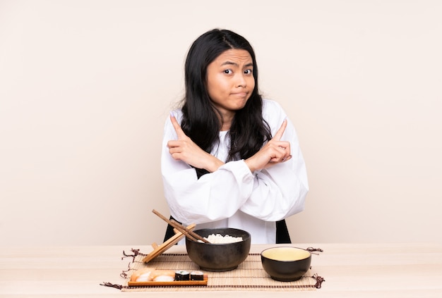 Adolescent fille asiatique manger de la nourriture asiatique isolé sur mur beige pointant vers les latéraux ayant des doutes