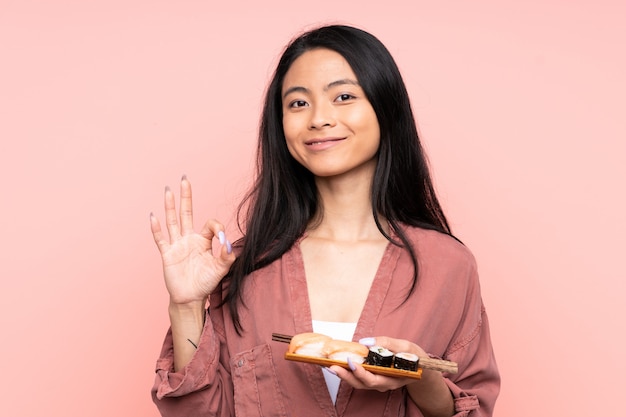 Adolescent fille asiatique manger des sushis isolé sur mur rose montrant un signe ok avec les doigts