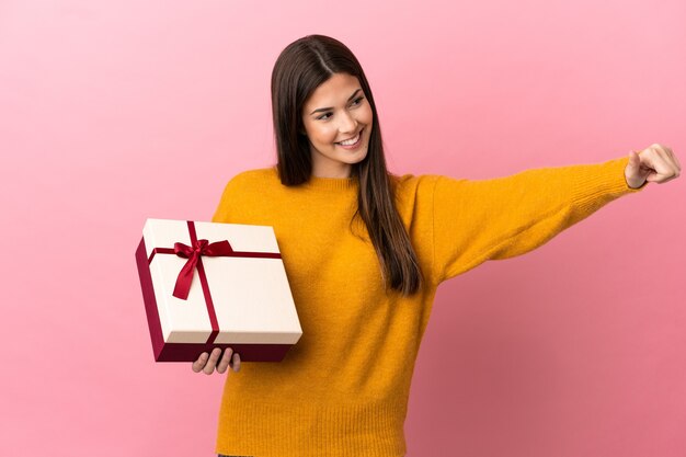 Adolescent fille brésilienne tenant un cadeau sur fond rose isolé donnant un geste de pouce en l'air