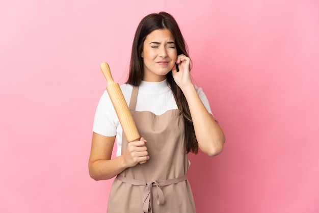 Adolescent fille brésilienne tenant un rouleau à pâtisserie isolé sur mur rose frustré et couvrant les oreilles