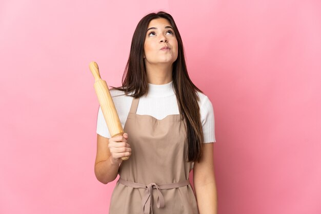 Adolescent fille brésilienne tenant un rouleau à pâtisserie isolé sur rose et levant