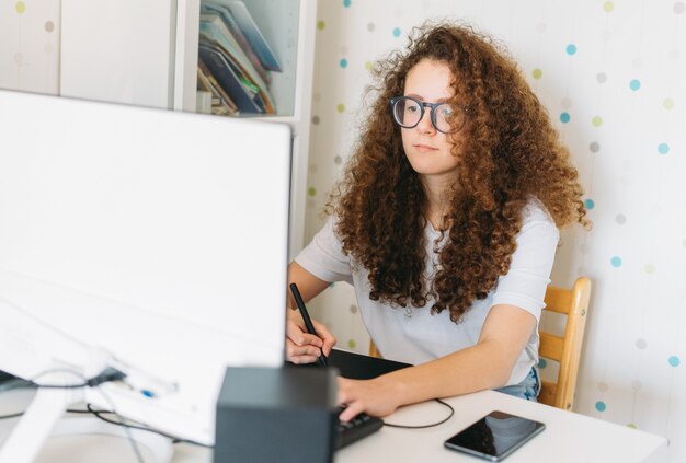 Adolescent fille cheveux bouclés yuong web designer dans des verres en t-shirt blanc assis travaillant avec tablette graphique sur ordinateur à la salle lumineuse à la maison