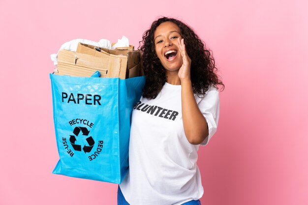 Adolescent fille cubaine tenant un sac de recyclage plein de papier à recycler isolé sur mur rose en criant avec la bouche grande ouverte
