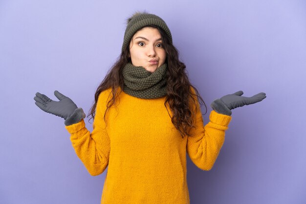 Adolescent fille russe avec chapeau d'hiver isolé sur mur violet ayant des doutes tout en levant les mains