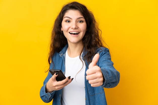 Adolescent fille russe isolée sur mur jaune à l'aide de téléphone mobile tout en faisant les pouces vers le haut