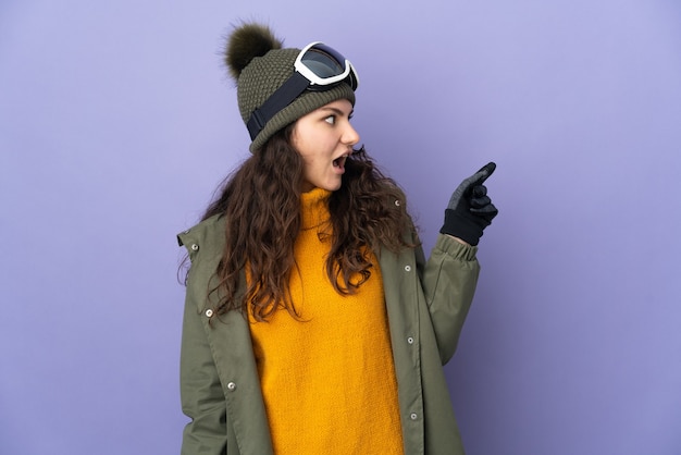 Adolescent fille russe avec des lunettes de snowboard isolé sur fond violet dans l'intention de réaliser la solution tout en levant un doigt
