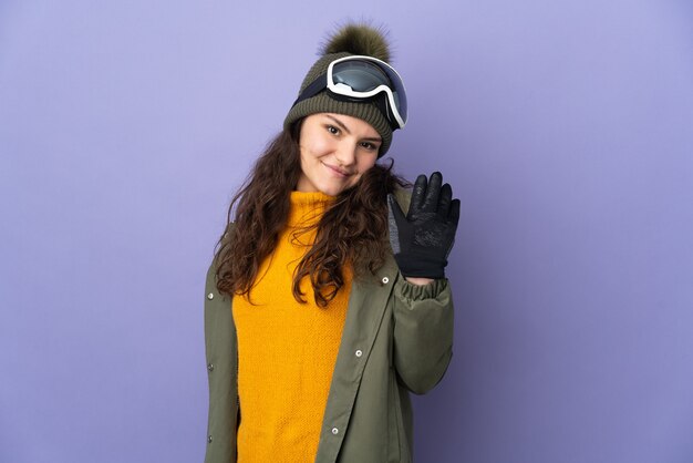 Adolescent fille russe avec des lunettes de snowboard isolé sur fond violet saluant avec la main avec une expression heureuse