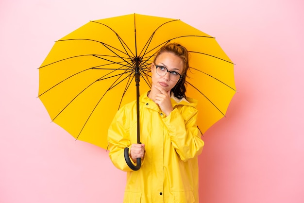 Adolescent fille russe avec manteau imperméable et parapluie isolé sur fond rose ayant des doutes