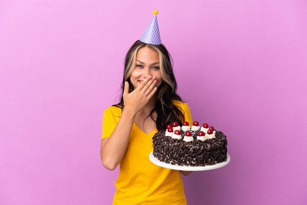 Adolescent fille russe tenant un gâteau d'anniversaire isolé sur fond violet heureux et souriant couvrant la bouche avec la main
