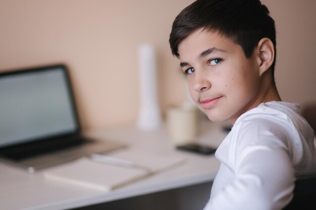 Un adolescent heureux est assis près de la table et étudie à la maison Un garçon étudie en ligne pendant la quarantaine Contexte