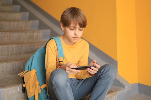 Un adolescent heureux vêtu d'un T-shirt jaune est assis avec un téléphone près d'un mur jaune. Utiliser la technologie en ligne