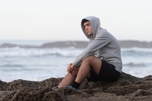 Adolescent hispanique inquiet assis sur des rochers au bord de la mer. Anxiété et dépression sur le concept de l'adolescence.