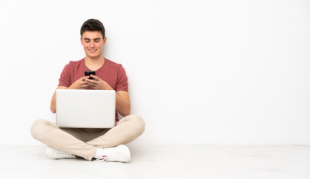 Adolescent homme assis sur le sol avec son ordinateur portable en envoyant un message avec le mobile