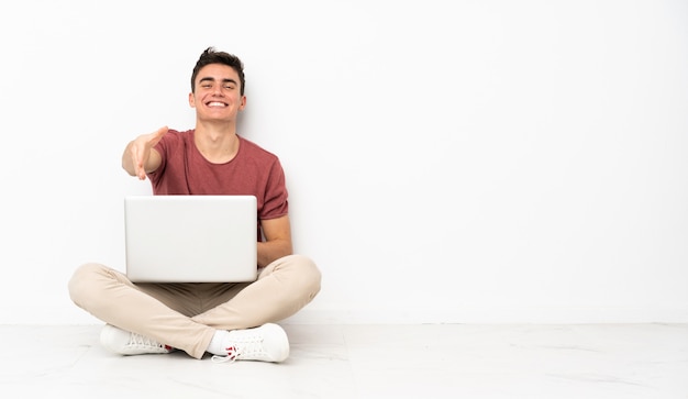 Adolescent homme assis sur le sol avec son ordinateur portable se serrant la main pour fermer une bonne affaire
