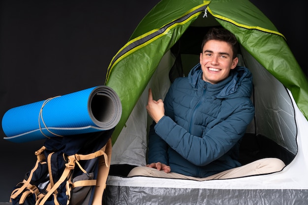 Adolescent homme caucasien à l'intérieur d'une tente de camping vert isolé sur fond noir pointant vers l'arrière