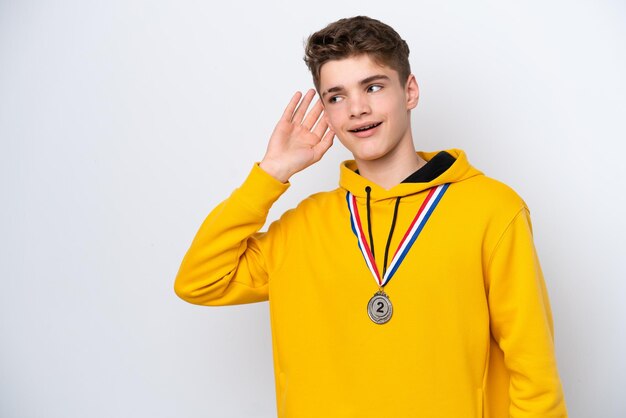 Adolescent homme russe avec des médailles isolé sur fond blanc écoutant quelque chose en mettant la main sur l'oreille