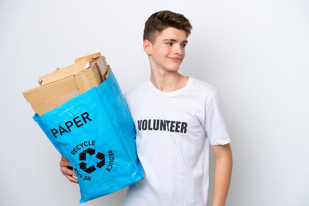Adolescent homme russe tenant un sac de recyclage plein de papier à recycler isolé sur fond blanc regardant sur le côté et souriant